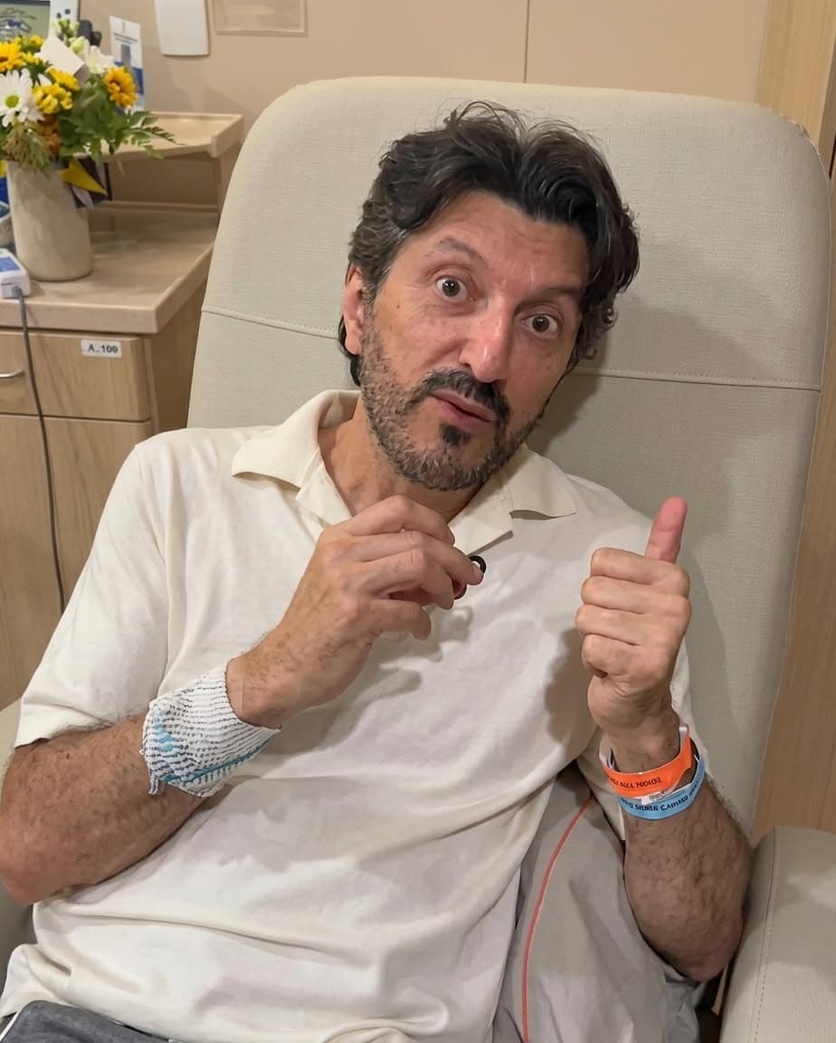Lito Sousa seated in a hospital recliner, wearing a light polo shirt and a hospital wristband, giving a thumbs up with flowers on a counter behind him.
